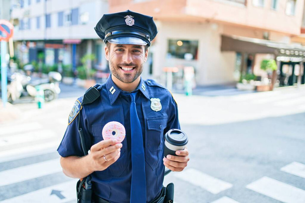 Νέος όμορφος Ισπανός αστυνομικός που φοράει αστυνομική στολή χαμογελώντας χαρούμενος. Τρώγοντας ντόνατ και πίνοντας φλιτζάνι καφέ take away στο δρόμο της πόλης. - Φωτογραφία, εικόνα