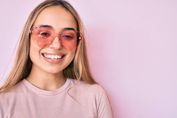 Young beautiful blonde woman wearing heart shaped sunglasses smiling with a happy and cool smile on face. showing teeth.  - Photo, Image
