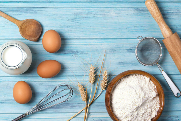 Ingredienti per cottura al forno, latte, uova, farina di frumento e stoviglie su fondo blu in legno, vista dall'alto - Foto, immagini