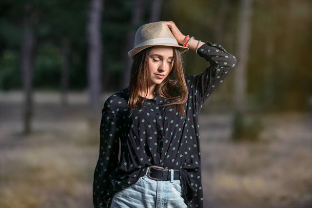 Una mujer joven vestida con una camisa de lunares con un sombrero de paja triste un día soleado sobre un fondo verde bosque
. - Foto, imagen