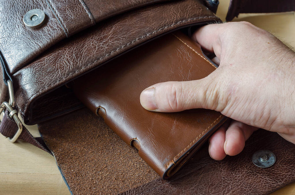 Male hand removes brown wallet from leather bag. Caucasian man takes out leather purse from an open bag. Shooting from behind. Selective focus. - Foto, Imagen