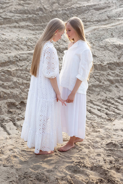 dwie młode siostry bliźniaczki pozujące na pustyni lub kamieniołomie piasku w eleganckich białych ubraniach - Zdjęcie, obraz