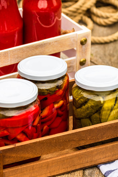 Wooden crate with glass jars with pickled red bell peppers and pickled cucumbers (pickles) isolated. Jars with variety of pickled vegetables. Preserved food concept in a rustic composition. - Photo, Image