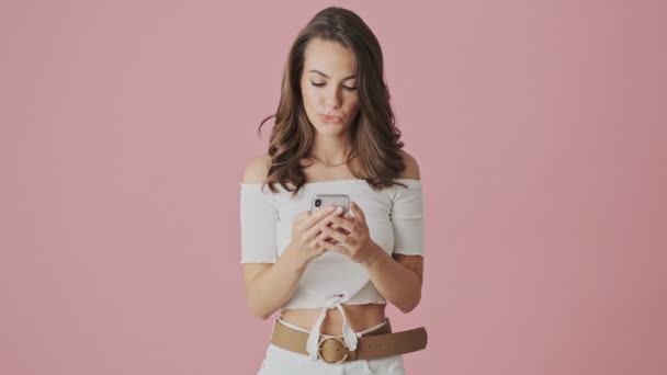 Egy boldog fiatal nő fantasztikus ajánlatot kap az okostelefonjára, amint elszigetelten áll rózsaszín háttérrel a stúdióban. - Felvétel, videó