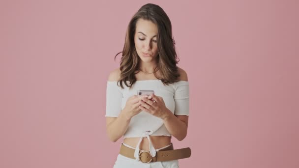 An emotional young woman is watching something amazing on her smartphone standing isolated over pink background in studio - Video