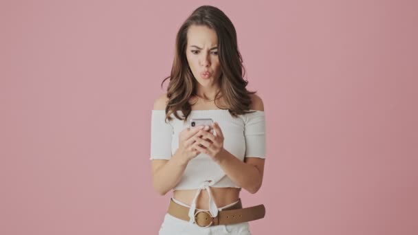 An angry young woman is typing on her smartphone standing isolated over pink background in studio - Πλάνα, βίντεο