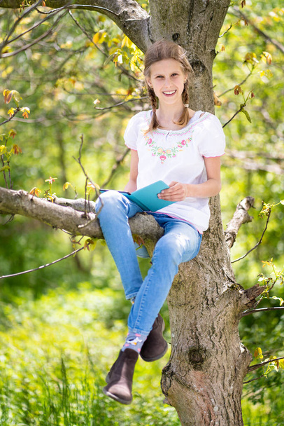 Ragazza sorridente adolescente in bianco T-shirt e jeans seduti su un albero, leggere un libro su ebook reader. Ottima idea e attività per trascorrere le vacanze estive nella natura. - Foto, immagini