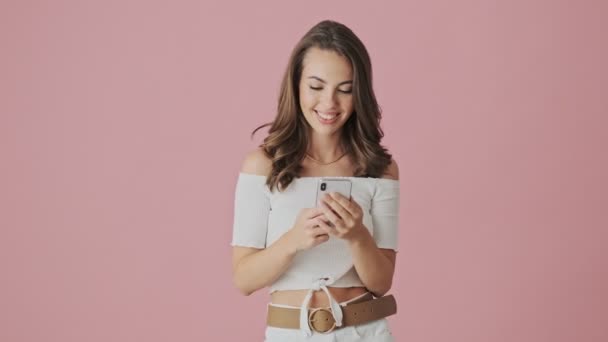 A pleased young woman is showing thumb-up gesture using her smartphone standing isolated over a pink background in studio - Filmati, video