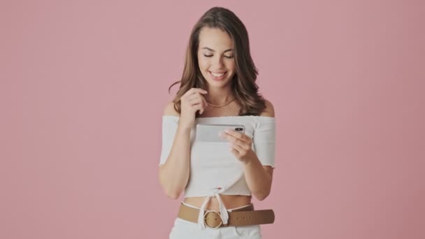 A cheerful young woman is watching something funny on her smartphone holding it horizontally standing isolated over a pink background in studio - Video