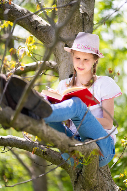 Tini lány fehér pólóban és rózsaszín kalapban ül egy fán a kertben, mosolyog és érdekes könyvet olvas. Nagyszerű ötlet és tevékenység tölteni nyári szünidő a természetben. - Fotó, kép