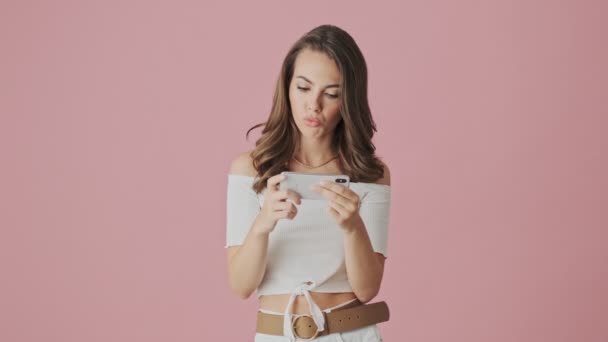 A focused young woman is watching something interesting on her smartphone holding it horizontally standing isolated over a pink background in studio - Séquence, vidéo