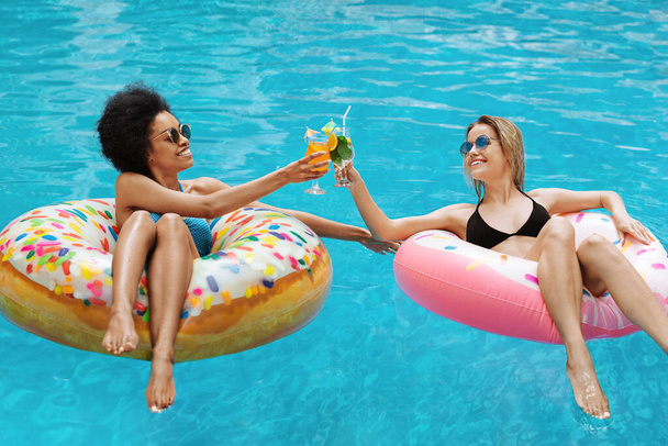 Ładne dziewczyny dobrze się bawią i piją zimne koktajle na nadmuchiwanych pierścieniach na basenie - Zdjęcie, obraz