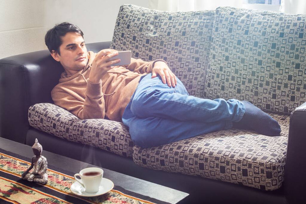 A young Hispanic male sitting on the couch and talking on the phone in the quarantine due to Covid-19 pandemic - the new normal concept - Photo, image