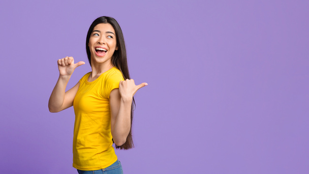 Joyful asian girl pointing on copy space behind her back with thumbs - Photo, Image