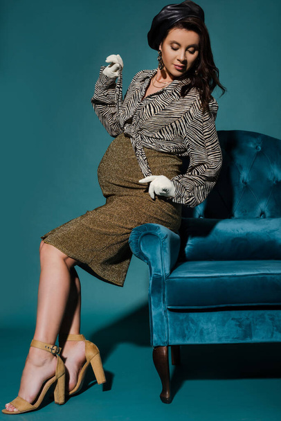 элегантная беременная женщина трогает полосатую блузку, позируя на велюровом кресле на бирюзовом - Фото, изображение
