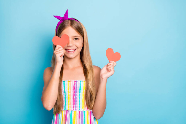 Portrait of positive cheerful kid girl close cover face with small red paper card heart enjoy 14-february holiday date event wear colorful dress skirt isolated over blue color background - 写真・画像