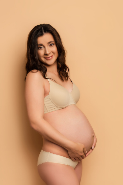 zwanger brunette vrouw in lingerie kijken naar camera terwijl aanraken buik op beige - Foto, afbeelding