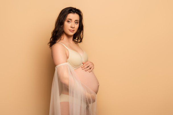 pregnant woman in lingerie and chiffon sleeves touching belly and looking at camera on beige - Photo, Image