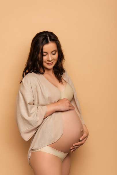 joven embarazada en lencería y camisa tocando barriga en beige - Foto, Imagen