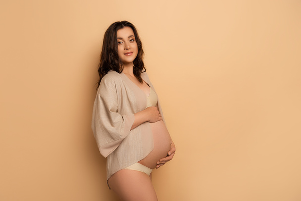 вагітна жінка в сорочці і нижній білизні дивиться на камеру, торкаючись живота на бежевому
 - Фото, зображення