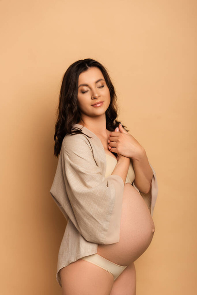 mujer embarazada soñadora en lencería y camisa sosteniendo las manos apretadas cerca del pecho en beige - Foto, Imagen