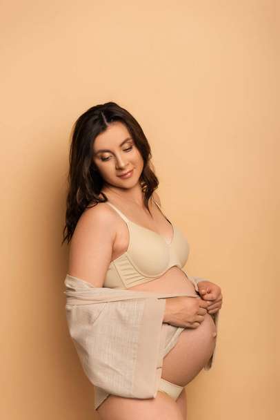 young pregnant woman in underwear covering belly with shirt on beige - Photo, Image
