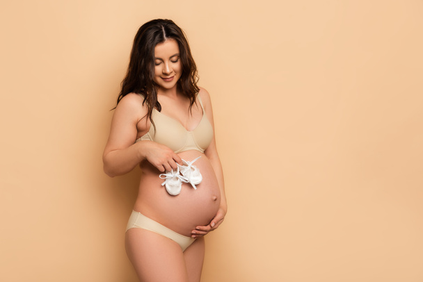 brunette pregnant woman in underwear touching belly and holding baby booties on beige - Foto, afbeelding
