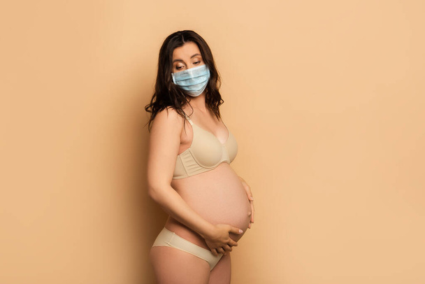 jonge zwangere vrouw in lingerie en beschermende masker aanraken buik en kijken neer op beige - Foto, afbeelding