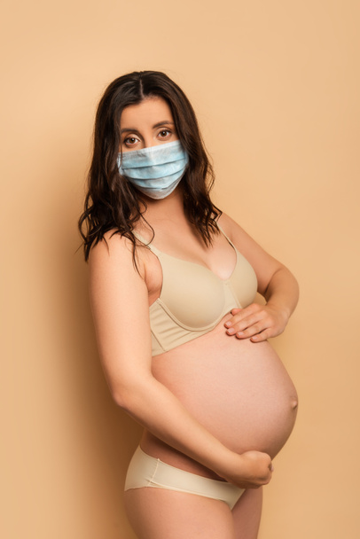 беременная женщина в нижнем белье и медицинской маске трогает животик и смотрит в камеру на бежевом - Фото, изображение