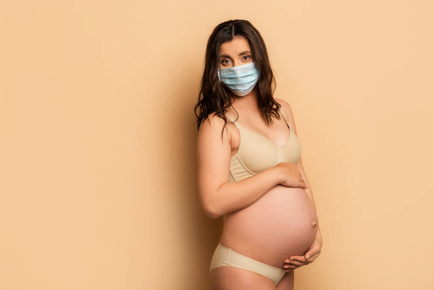 jovem grávida em roupa interior e máscara médica tocando barriga e olhando para a câmera no bege
 - Foto, Imagem