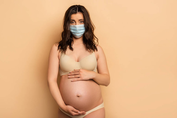 μελαχρινή, έγκυος γυναίκα με προστατευτική μάσκα που αγγίζει την κοιλιά και κοιτάζει την κάμερα σε μπεζ - Φωτογραφία, εικόνα