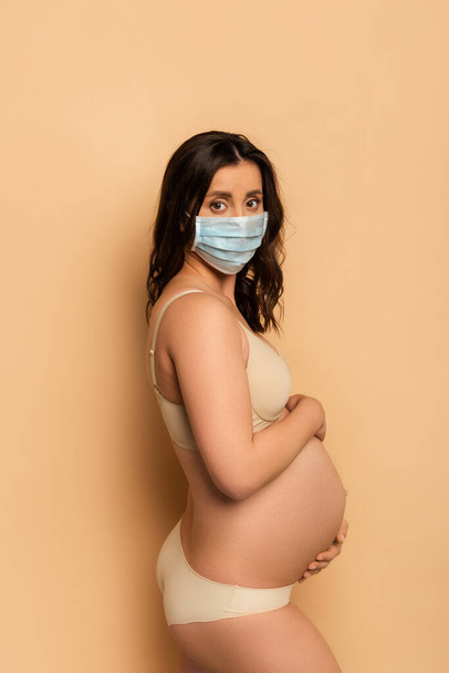 morena, mujer embarazada en ropa interior y máscara médica tocando la barriga y mirando a la cámara en beige - Foto, imagen