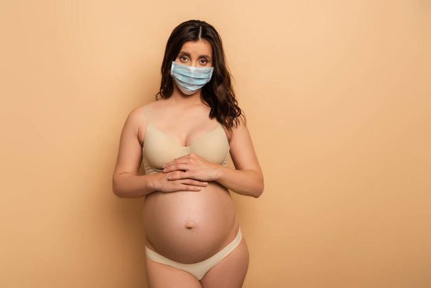 беременная женщина в нижнем белье и медицинской маске смотрит в камеру и трогает живот на бежевый - Фото, изображение