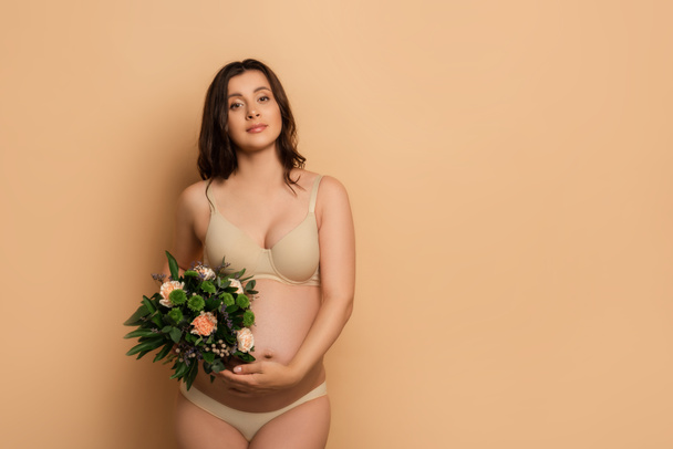 young pregnant woman in lingerie holding bouquet while looking at camera on beige - Photo, Image