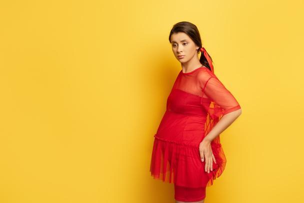 mujer embarazada preocupada en túnica roja tocando la cadera mientras está de pie en amarillo - Foto, imagen