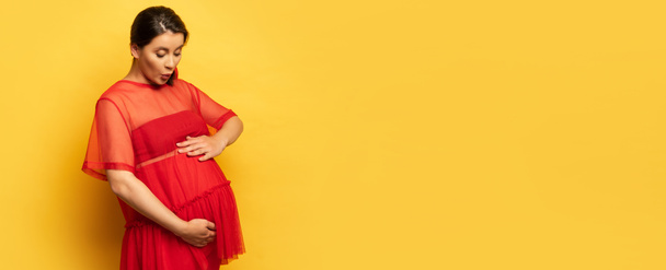 encabezado del sitio web de conmocionado, mujer joven embarazada en túnica roja tocando la barriga en amarillo - Foto, Imagen