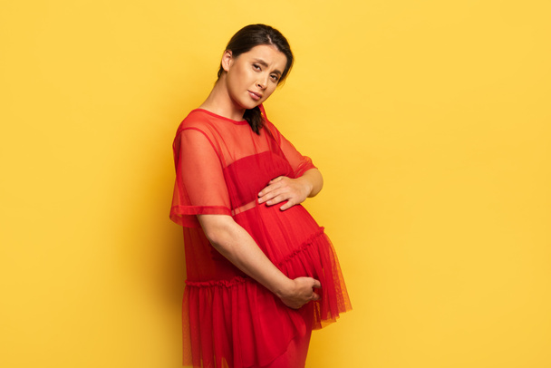 έγκυος νεαρή γυναίκα σε κόκκινο στολή ανησυχούν ενώ αγγίζει την κοιλιά και κοιτάζοντας κάμερα σε κίτρινο - Φωτογραφία, εικόνα