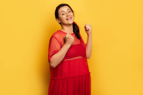 excited pregnant woman in red outfit showing winner gesture while looking at camera on yellow - Фото, изображение