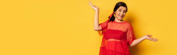 panoramic shot of pregnant woman in red tunic standing with open arms while looking at camera on yellow - Photo, Image