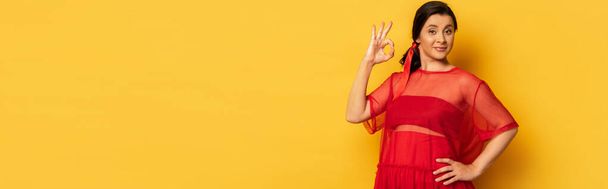 горизонтальное изображение беременной женщины в красной тунике, показывающей нормальный жест, держа руку на бедре на желтом - Фото, изображение