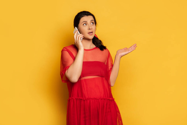 mujer embarazada confundida en traje rojo que muestra gesto de encogimiento mientras habla en el teléfono inteligente en amarillo - Foto, imagen