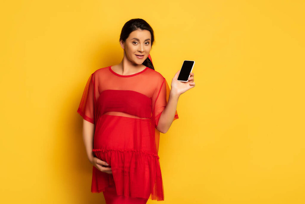 bruna donna incinta in tunica rossa che mostra smartphone con schermo bianco mentre tocca la pancia sul giallo - Foto, immagini