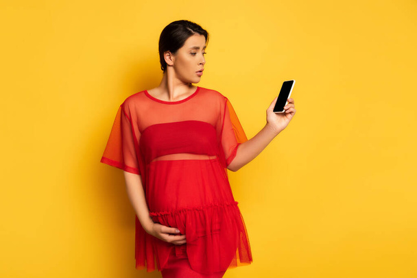 Verärgerte schwangere Frau in roter Tunika hält Smartphone mit leerem Bildschirm, während sie Bauch auf gelb berührt - Foto, Bild