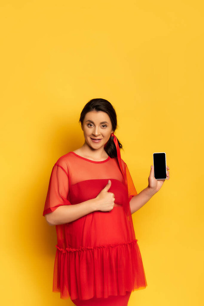 Kırmızı elbiseli hamile kadın, sarı ekranda boş ekranlı akıllı telefonu tutarken baş parmağını gösteriyor. - Fotoğraf, Görsel