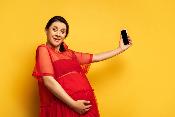 brünette schwangere Frau in roter Tunika macht Selfie auf dem Smartphone, während sie in die Kamera auf gelb schaut - Foto, Bild