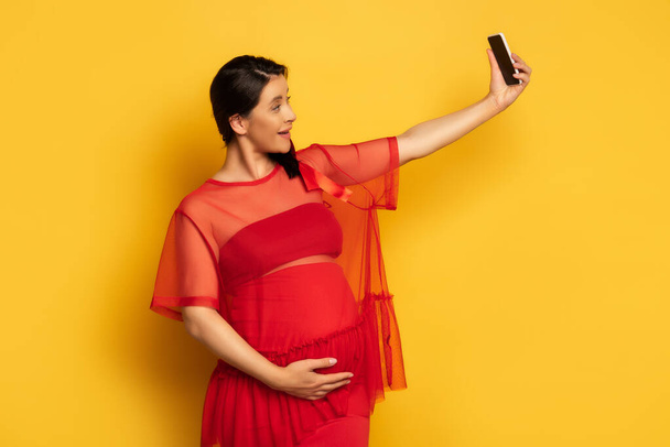 mujer embarazada en túnica roja tocando la barriga mientras toma selfie en el teléfono inteligente en amarillo - Foto, imagen