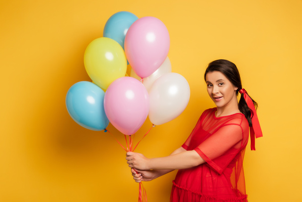 brunette pregnant woman in red outfit looking at camera while holding colorful festive balloons on yellow - Photo, Image