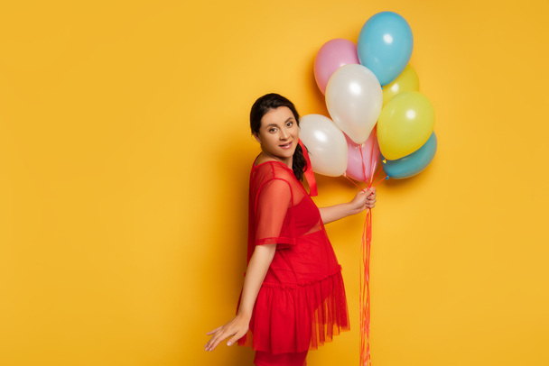 pregnant woman in red tunic holding colorful festive balloons while looking at camera on yellow - Photo, Image
