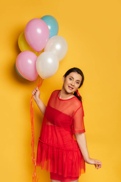pregnant woman holding multicolored festive balloons while looking at camera on yellow - Foto, Imagem