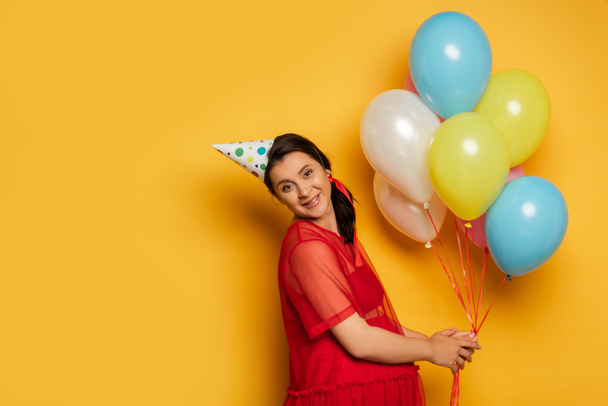 mujer embarazada con gorra de fiesta sosteniendo globos festivos multicolores en amarillo   - Foto, Imagen
